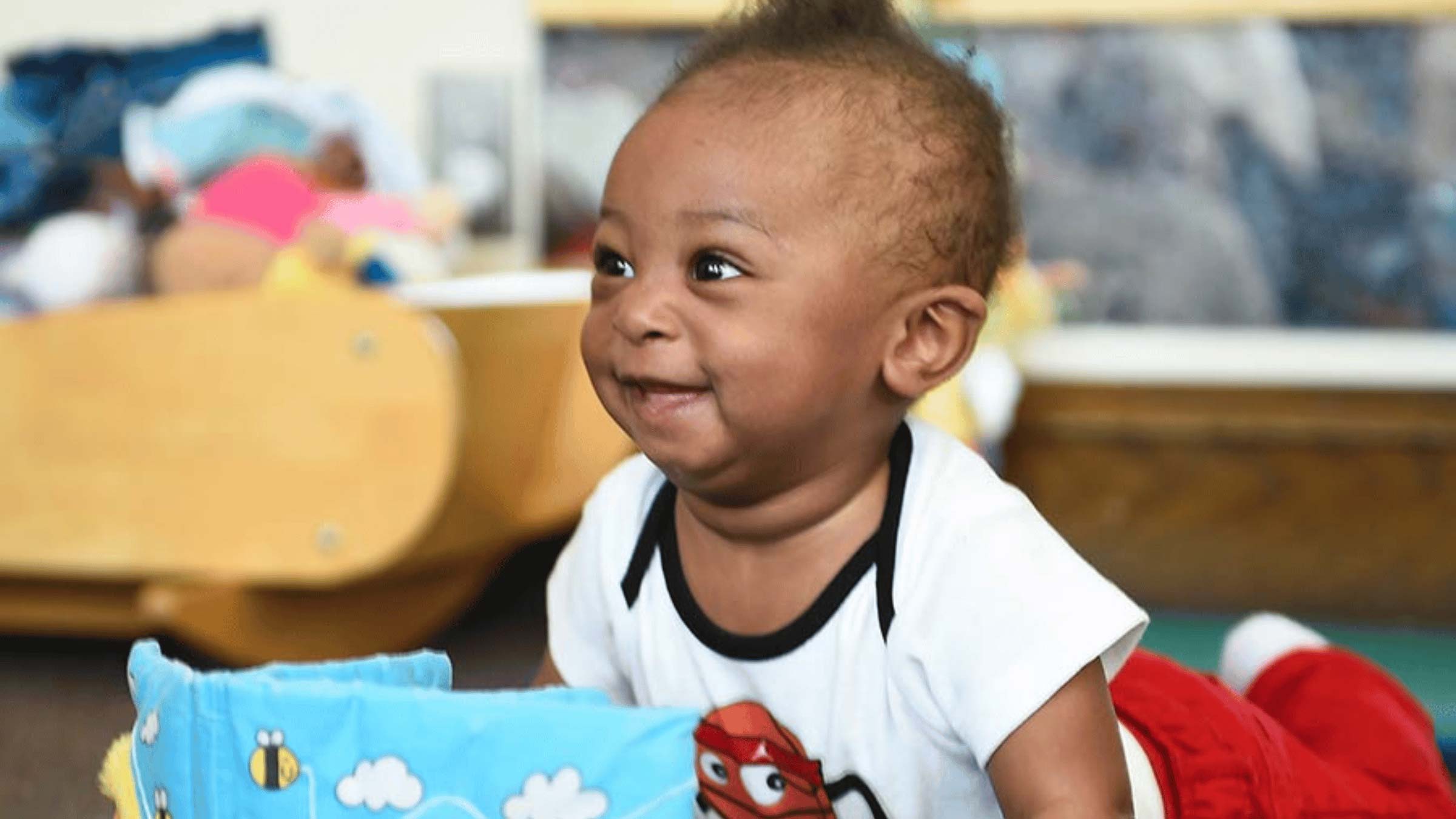 A baby sits on the floor with a book in front of them and smiles