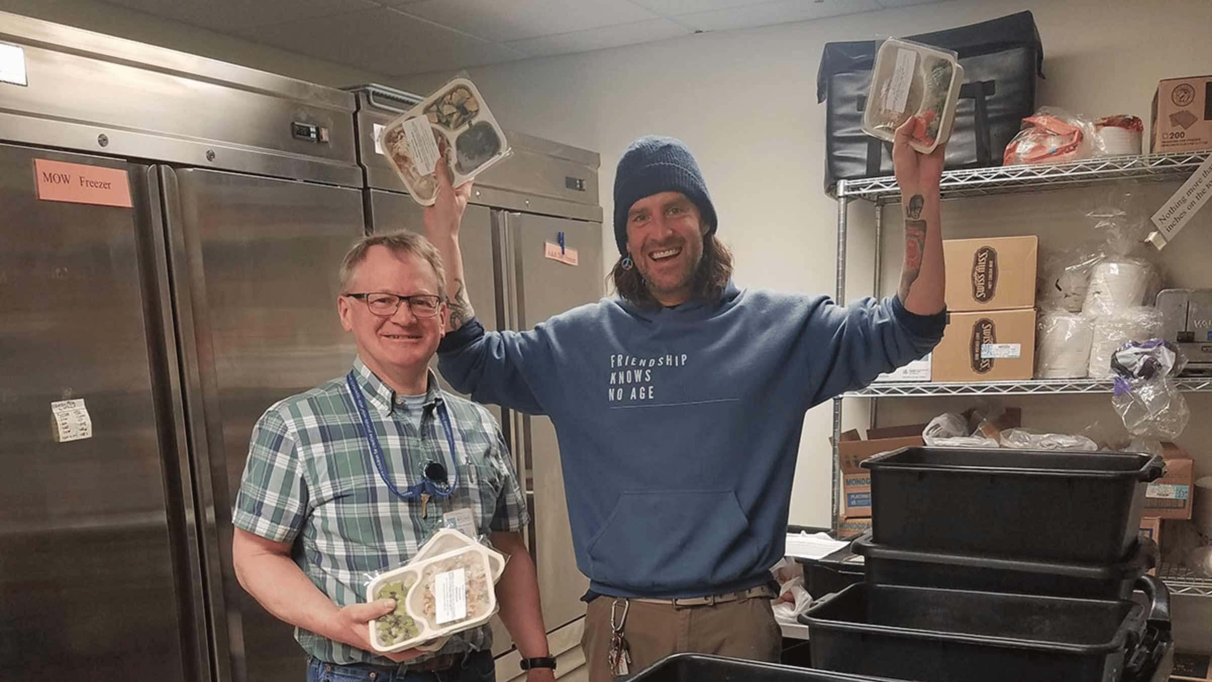 Two people stand in a kitchen holding up meal packages and smiling