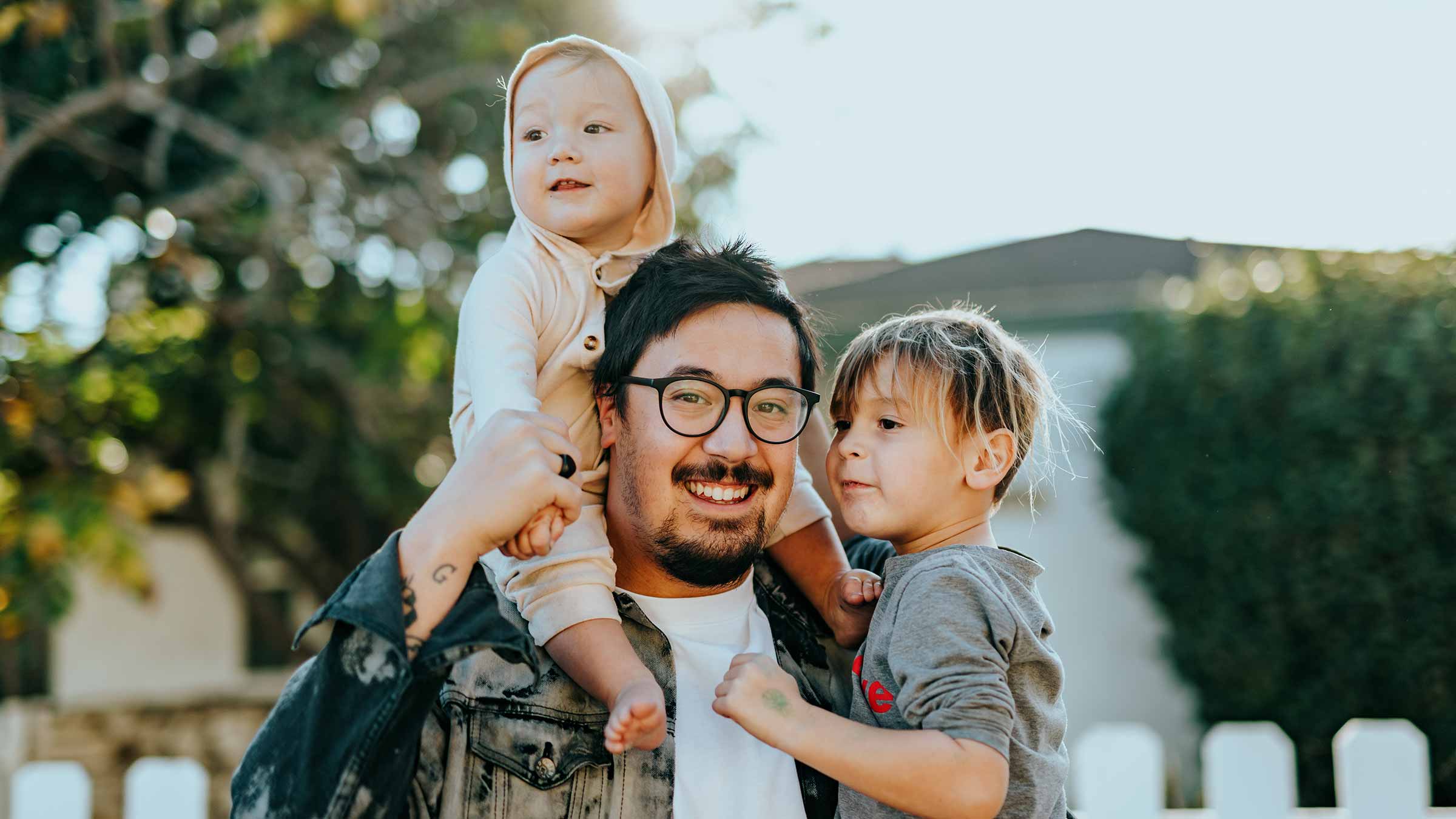 A father stands holding one child and the other sits on his shoulders