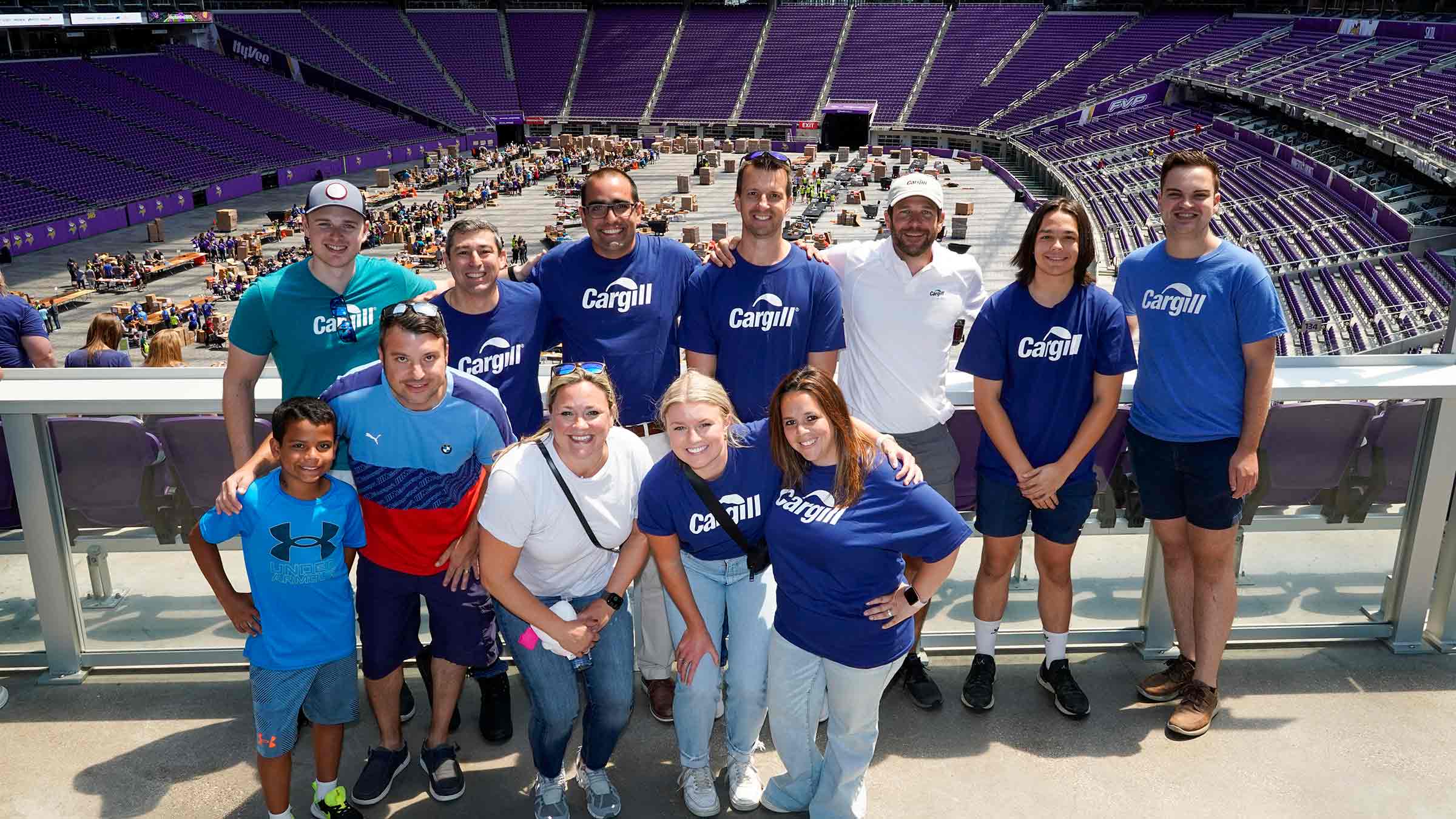 A group of Cargill volunteers stand together at US Bank Stadium while Action Day volunteers are packing school supplies behind them