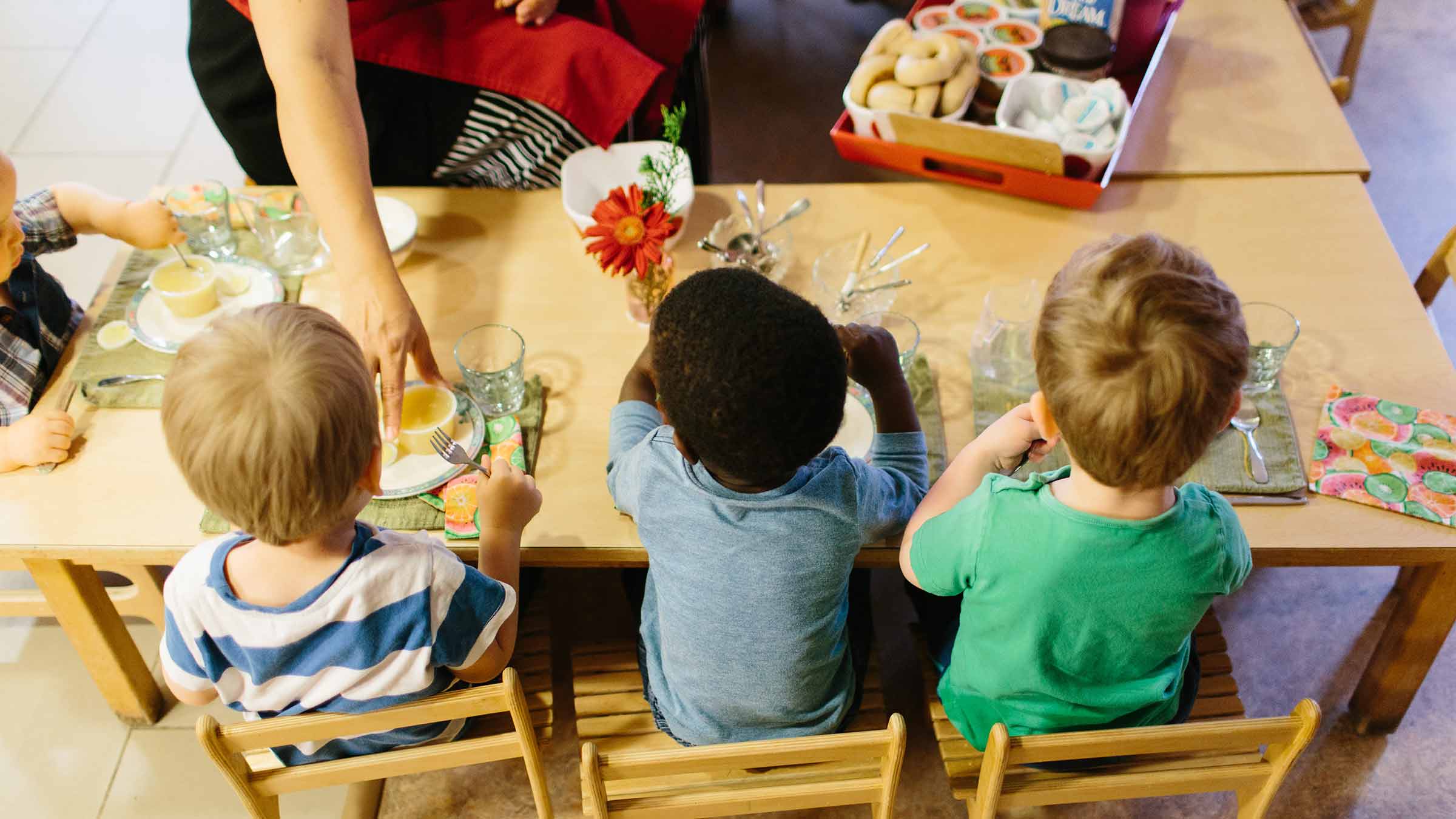 A group of preschoolers sit at a table together while their teacher hands out their snack