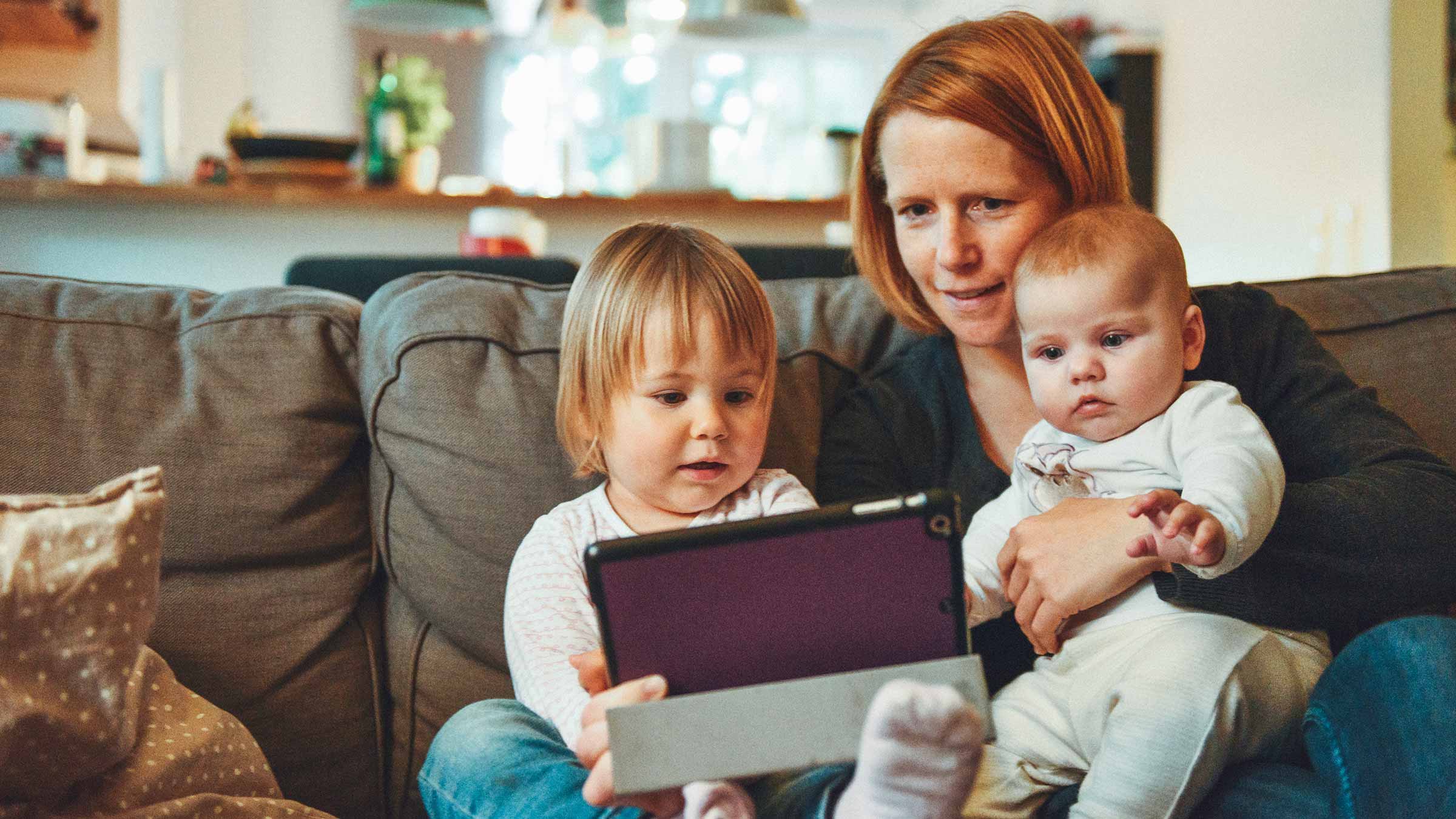 A mother sits on the couch with her two young children looking at an iPad