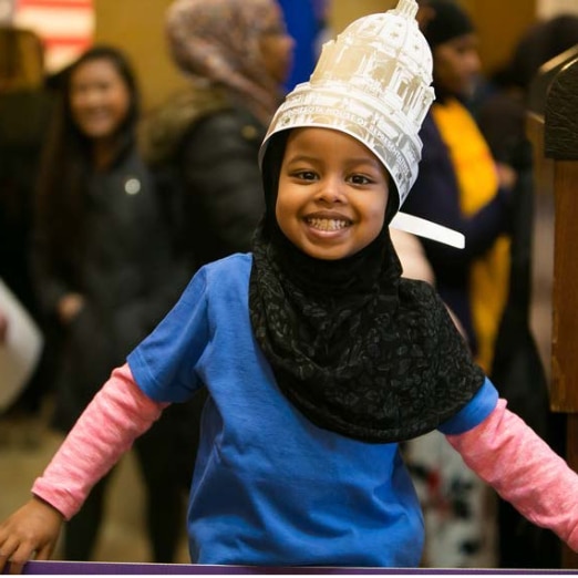 A young child smiles at the camera while wearing a hat that looks like the Minnesota state capital building