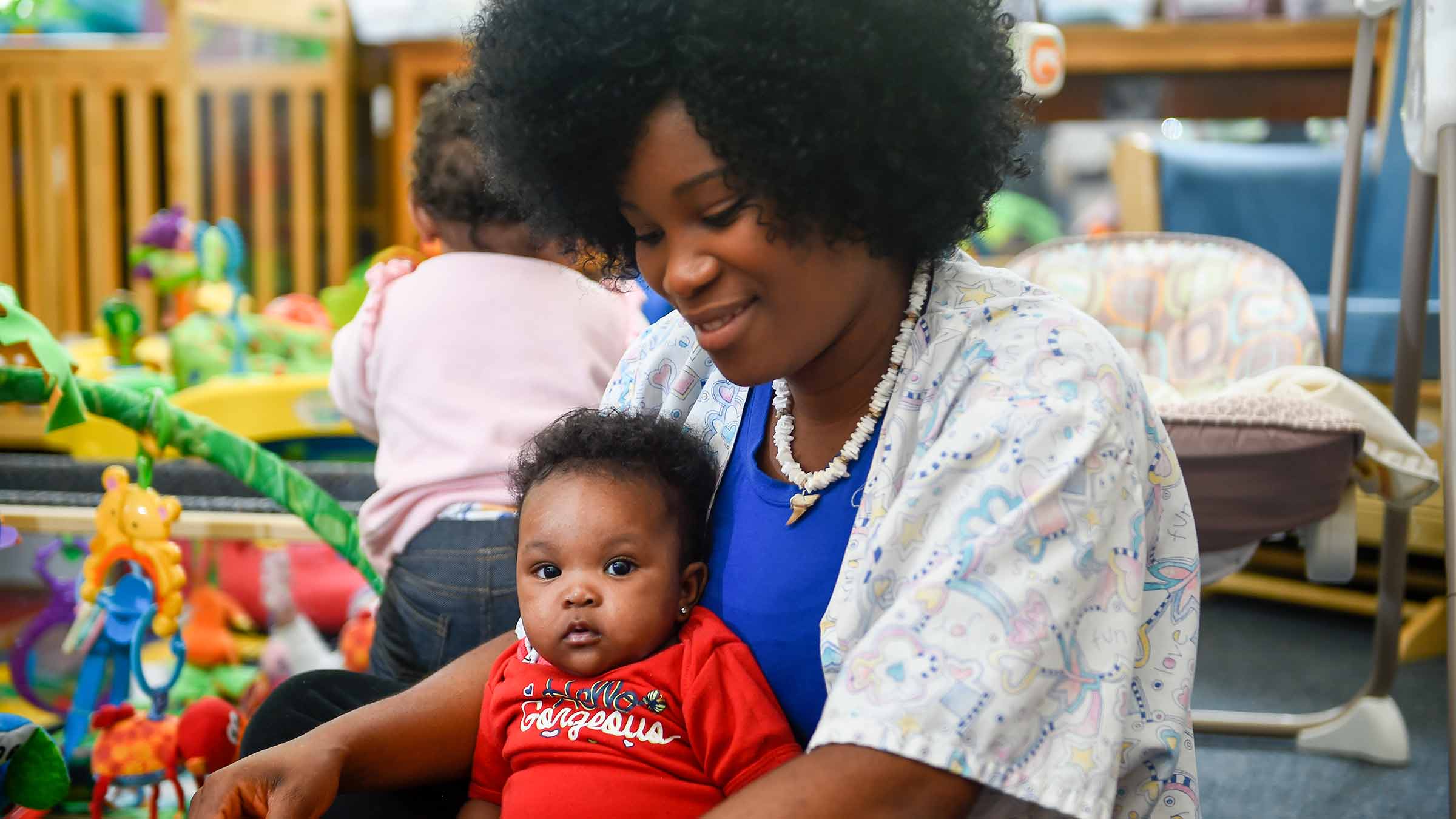 A mother sits in a preschool room with her baby on her lap