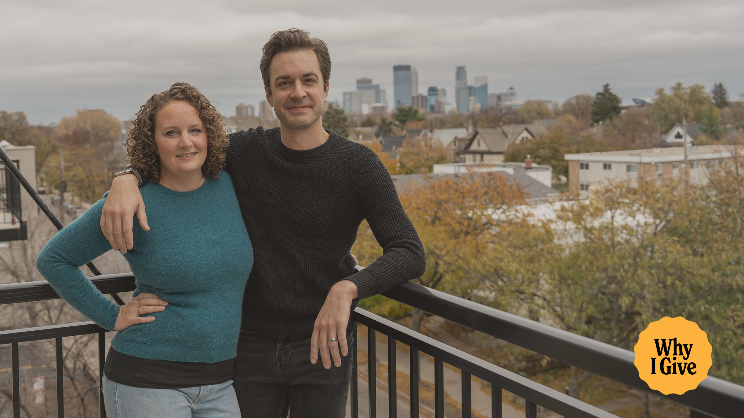 A couple smiles outside on a balcony with a city in the background