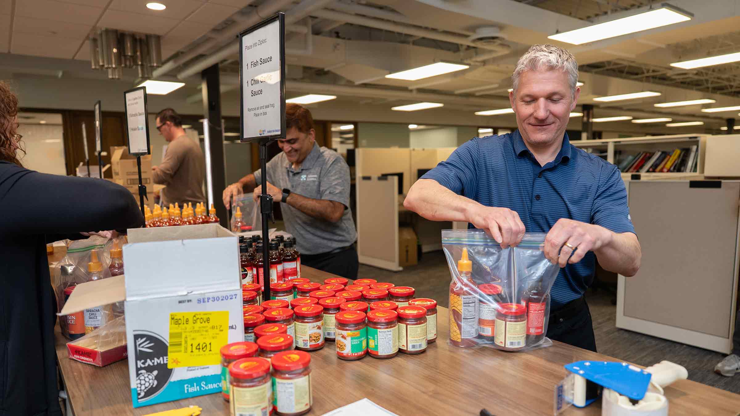 Volunteers assemble food kits with sauces and packaged items at an indoor distribution table.